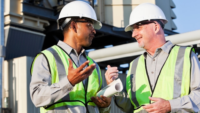 Two men wearing hardhats, safety glasses, and high visibility vests talking