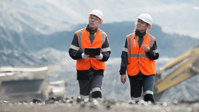 Two people walking on a mining site wearing safety glasses, hard hats, and high visibility vests