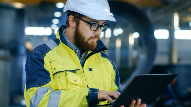 Man in hardhat and construction jacket typing on laptop in production facility