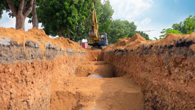 A trench with a man standing in it and an excavator at the far side.