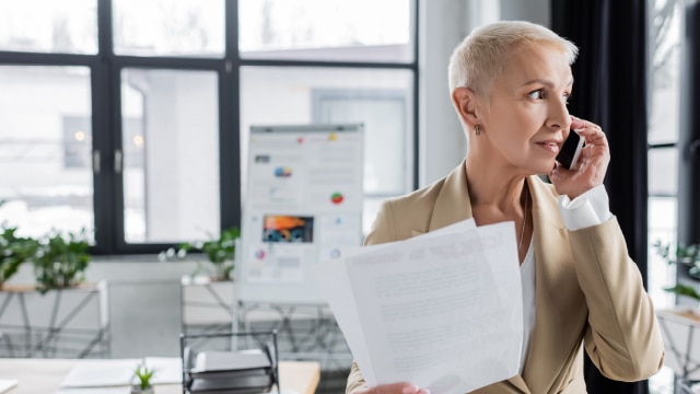 Woman looking out of her office window while holding two pieces of paper and talking on the phone