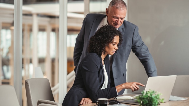 Man standing behind woman and looking at laptop screen