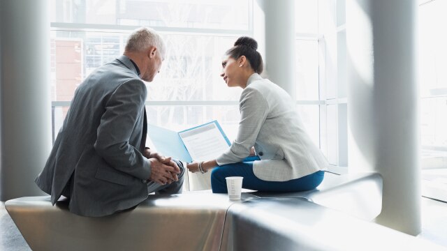 Two business people sitting on a bench and talking while holding a laptop