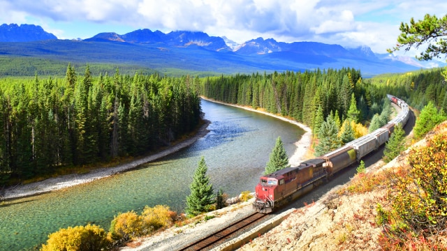 A train driving through the forest next to a river with mountains in the background