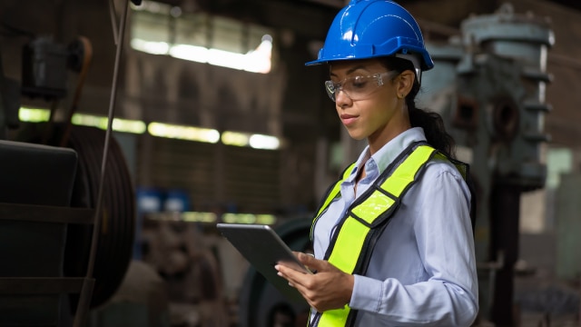 Woman wearing high visibility vest and hardhat looking at electronic tablet while in manufacturing facility.